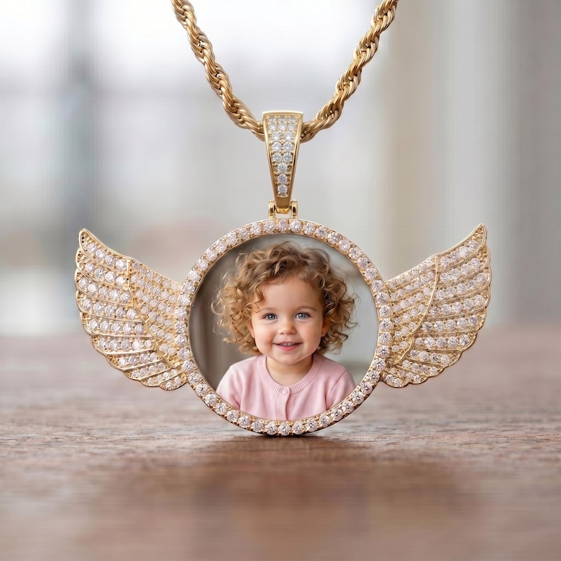 May include: A gold-toned circular pendant necklace featuring a central photograph of a curly-haired child wearing a pink shirt. The pendant is framed by clear stones and adorned with two symmetrical, stone-encrusted angel wings on each side.
