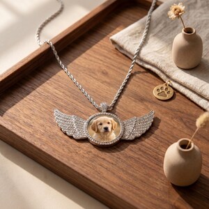 May include: A silver-toned pendant necklace featuring a circular frame with a photograph of a golden retriever puppy, flanked by two crystal-encrusted angel wings. The pendant hangs from a twisted silver chain, accompanied by a small paw print charm.