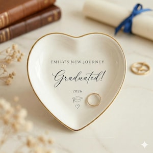May include: A heart-shaped, white ceramic trinket dish with a gold rim. The dish is inscribed with "EMILY'S NEW JOURNEY Graduated! 2026" and features a graduation cap and heart icon. A gold ring rests inside the dish.