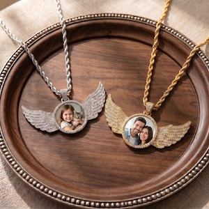 May include: Two circular pendant necklaces with rope chains, one silver-toned and one gold-toned. Each pendant features a central circular photo frame surrounded by textured angel wings. The silver pendant displays a child with a cat, while the gold pendant shows a couple.