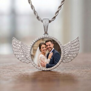 May include: A silver-toned circular pendant necklace featuring a photo of a couple in wedding attire. The pendant is framed by small clear stones and adorned with two large, stone-encrusted angel wings on either side, hanging from a twisted chain.