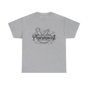 Puede incluir: Camiseta gris con un gráfico negro de un mortero y una maja con la palabra "Pharmacist" en una fuente cursiva. El diseño incluye ilustraciones de pastillas alrededor del mortero y la maja.