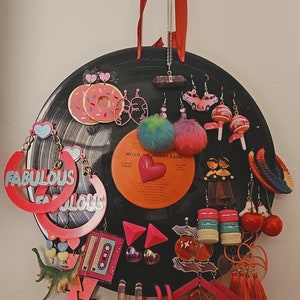 May include: A black vinyl record with a variety of colorful earrings hanging from it. The earrings include hearts, donuts, cherries, a cassette tape, a lightning bolt, a dinosaur, a pom-pom, and a hexagonal design. The record has a label with the text "HELLO I'M JOHNNY CASH" on it.