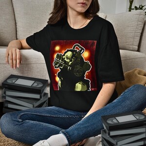 May include: A black short-sleeve t-shirt featuring a graphic print of a pale, masked figure wearing a mechanical head device with gears and wires against a dark red background with glowing yellow circular lights.