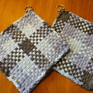 May include: Two woven pot holders with a checkered pattern in shades of blue and gray. Each pot holder has a metal ring attached to the top.