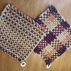 May include: Two woven pot holders with a brown, beige, and burgundy checkered pattern. The pot holders have a white loop attached to each corner.