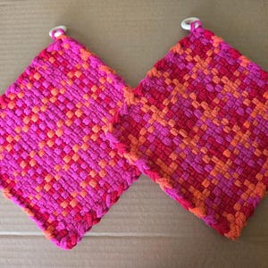 May include: Two pink and orange woven pot holders with white loops. The pot holders are made of a thick, textured yarn.