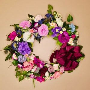 May include: A vibrant floral wreath featuring an assortment of colorful flowers, including pink roses, purple hydrangeas, and white blossoms. A large burgundy bow adds a decorative touch. Green leaves are interspersed throughout the arrangement.