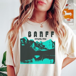 May include: Cream-colored t-shirt featuring a graphic of Banff National Park. The design includes the word "BANFF" in bold, dark green letters above a teal and dark green mountain and lake scene. The words "NATIONAL PARK" are below the mountain graphic.