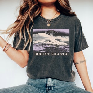 May include: A dark gray t-shirt with a graphic print of a mountain with a UFO flying over it. The text "MOUNT SHASTA" is printed below the graphic.