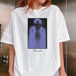 Puede incluir: Camiseta blanca con un estampado gráfico de una persona con un atuendo estampado en azul y blanco. El gráfico está sobre un fondo negro y el texto "Metropolis" está impreso debajo.