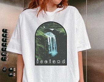 May include: White t-shirt with a graphic of a waterfall in a green landscape. The graphic is surrounded by a black circle and the word "Iceland" is printed below the graphic.