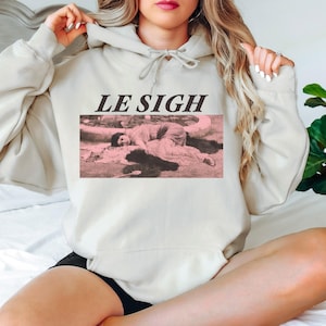 May include: A beige hoodie with the text "LE SIGH" above a pink and black image of a person lying down. The hoodie has a drawstring hood and a kangaroo pocket.