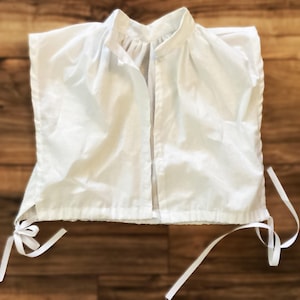May include: White cotton sleeveless top with a high collar and a button closure at the front. The top has a gathered detail at the neckline and ties at the bottom.