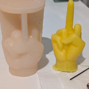 May include: A light peach-colored silicone mold and a bright yellow wax candle, both shaped like a hand making a middle finger gesture. The mold is cylindrical with a flat top, while the candle is a solid, detailed wax casting of the hand.