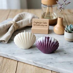 May include: Two shell-shaped candles, one solid cream and one ombre purple and white, sit on a marble surface. A wooden block displays the text Crafted for Calm. QtCandles.etsy.com. The candles feature a ribbed texture and a single centred wick.
