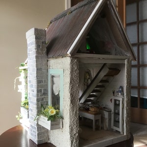 May include: A miniature dollhouse with a white exterior, a brown metal roof, and a gray stone chimney. The dollhouse has a window with a blue and white curtain, a flower box with yellow flowers, and a small interior with a staircase, a kitchen, and a cabinet.