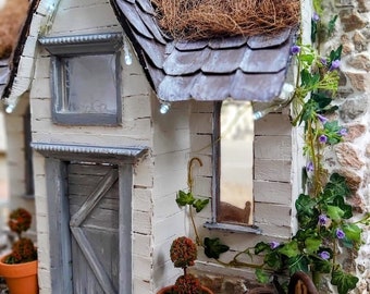 May include: A miniature white cottage with a thatched roof, a gray door, and two windows. The cottage is surrounded by greenery, including a small potted tree and a vine with purple flowers.