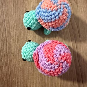 May include: Two handmade turtle toys. One turtle has a light green head and limbs with a shell of blue, pink, and orange rubber bands. The other turtle has a light green head and limbs with a shell of pink, purple, and red rubber bands.
