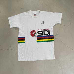 May include: White t-shirt featuring a colourful logo for Sidi, a cycling shoe brand. The logo includes the text "The Original from Italy" and a rainbow stripe design. The shirt also has a small Ultima logo on the chest.