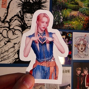 May include: A sticker featuring a person with pink hair, wearing a denim vest and jeans, forming a heart shape with their hands. The sticker has a white border and is held against a background of various images.