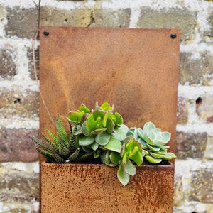 May include: A rectangular, rusted metal wall planter filled with various green succulents. The planter is mounted on a weathered brick wall. The succulents include a variety of textures and shades of green.