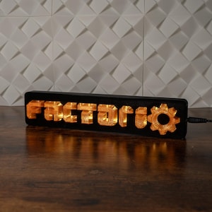 Puede incluir: Un letrero rectangular negro iluminado con la palabra "FACTORY" en letras naranjas y un símbolo de engranaje. El letrero está enchufado y descansa sobre una superficie de madera, con una pared blanca texturizada de fondo.