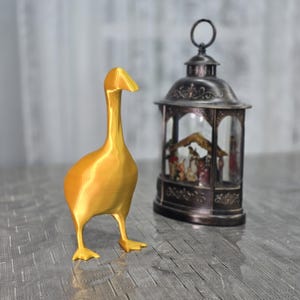 Peut inclure: Une figurine de canard en or avec un long cou et un design détaillé. La figurine est posée sur une surface grise à côté d'une lanterne en métal noir avec une enceinte en verre.