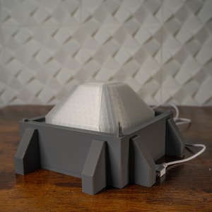 May include: A grey, geometric lamp with a white, translucent, pyramid-shaped shade. The lamp sits on a rectangular base with angled supports. A white cord and plug are attached to the base. The background features a white, textured wall.