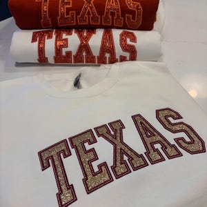 Può includere: Una pila di felpe in vari colori, tra cui bianco, rosso e turchese. Ogni felpa presenta la scritta "TEXAS" in un carattere glitterato in stile collegiale. Le felpe sono piegate ordinatamente, mettendo in mostra le scritte audaci.