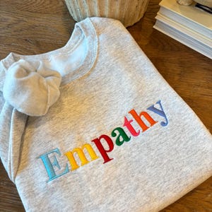 May include: A folded light grey sweatshirt embroidered with the word "Empathy" in rainbow-coloured letters. The collar and part of the sleeve are visible. The embroidery features blue, yellow, red, and green, creating a vibrant design on the garment.