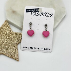 Könnte beinhalten: Ohrringe mit rosafarbenen Herzanhängern. Die Ohrringe sind auf einer weißen Karte mit dem Text "created by CROWS" und "HAND MADE WITH LOVE" abgebildet. Ein goldener Glitzerstern und eine weiße marmorähnliche Oberfläche befinden sich im Hintergrund.