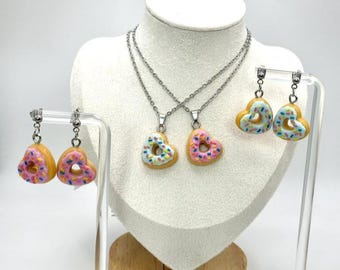 May include: A jewelry set featuring heart-shaped donut pendants and earrings. The donuts have a yellow base, pink or light blue frosting, and colorful sprinkles. The necklaces have silver chains, and the earrings have silver hooks.