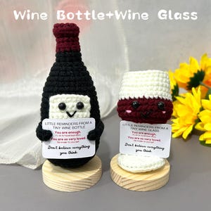Könnte beinhalten: Gehäkeltes Weinflaschen- und Weinglas-Set. Die Flasche ist schwarz mit einem bordeauxroten Oberteil, das Glas ist weiß und bordeauxrot. Jedes hat ein lächelndes Gesicht und ein kleines Schild mit Text. Auf Holzsockeln präsentiert.