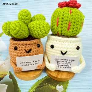 May include: Two crocheted potted plants with smiling faces and encouraging messages. The cactus plant is green with a red flower on top and the other plant has green leaves. Both plants are sitting on wooden bases.