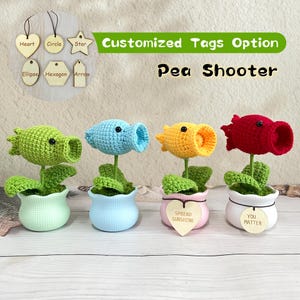 May include: Four crocheted pea shooter plants in green, blue, yellow, and red, each in a small pot. The plants have green leaves and are labeled "Pea Shooter". Wooden tags with shapes and text like "Spread Sunshine" are also visible.