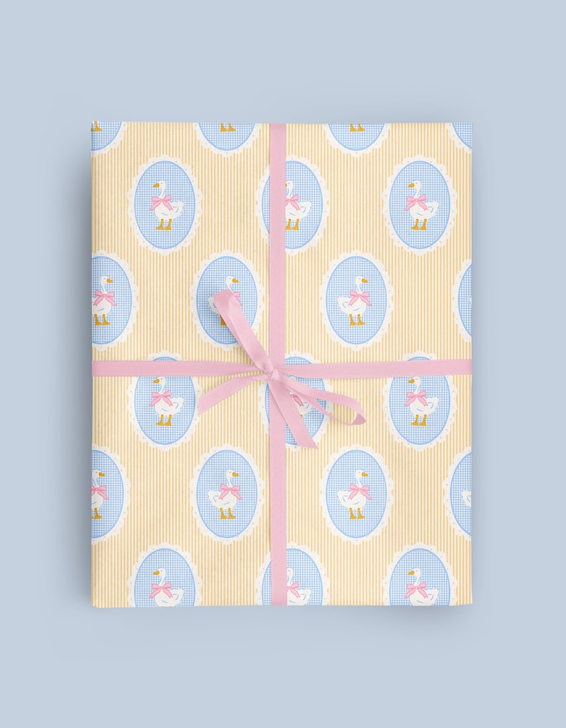 Puede incluir: Paquete de regalo envuelto en papel estampado con gansos en &oacute;valos azules con lazos rosas, sobre un fondo a rayas amarillas. El paquete est&aacute; atado con una cinta rosa.