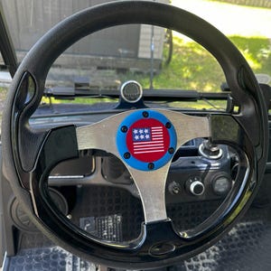 May include: Black steering wheel with a silver center and a patriotic emblem. The emblem features a red and white American flag design with a blue border and white stars. The steering wheel is part of a golf cart.