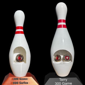 May include: Two white bowling pin-shaped display stands with red stripes. Each stand holds a ring with a red stone. One stand has the text "300 Game 800 Series" and the other reads "Terry 300 Game".