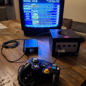 Może przedstawiać: Czarna konsola GameCube z czarnym kontrolerem i czarnym zasilaczem. Konsola jest podłączona do telewizora CRT wyświetlającego menu gier, w tym "Władca Pierścieni: Dwie Wieże", "TimeSplitters: Future Perfect" i "Tony Hawk's Pro Skater 3".