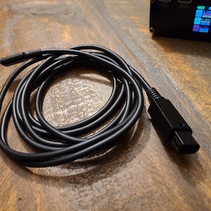 May include: A black cable with a USB-C connector on one end and a black connector on the other end. The cable is coiled up on a wooden surface.