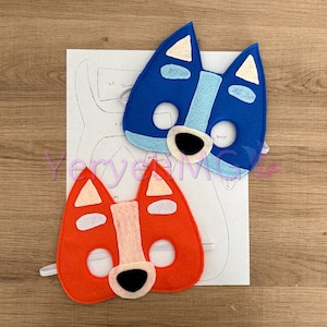 Puede incluir: Dos máscaras de animales de fieltro, una azul y otra naranja, con detalles blancos. Las máscaras tienen ojos y narices negros y están diseñadas para parecer perros.