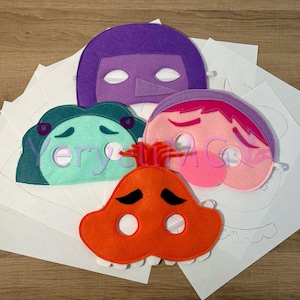 Puede incluir: Cuatro máscaras de fieltro con diferentes expresiones faciales. Las máscaras son de color morado, verde, rosa y naranja. Las máscaras están sobre un fondo blanco.