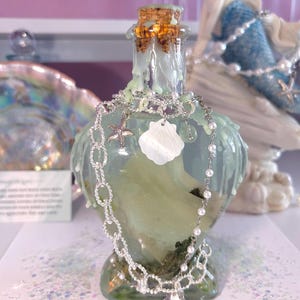 May include: A heart-shaped glass bottle with a cork stopper, covered in dripping light green wax. A silver-toned chain necklace featuring a small starfish charm, a white shell pendant, and a dangling silver seashell charm rests around the bottle neck.