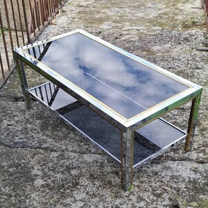 May include: A chrome and glass coffee table with a lower shelf. The table has a gold trim around the edges of the glass top.
