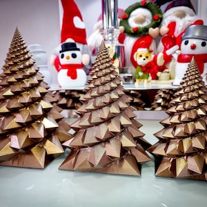 May include: Three gold-colored, geometric, Christmas tree ornaments. The trees are made of a series of triangular shapes that spiral upwards to form a cone shape.