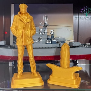 May include: A 3D printed gold-coloured figurine of a sailor wearing a peacoat and a sailor hat. The figurine is standing on a base. Behind the figurine is a 3D printed model of a battleship. The text "MR INNOVATIONS INNOVATING THE IMPOSSIBLE" is visible in the background.