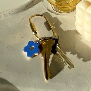 May include: A gold keychain with a blue flower charm and several keys rests on a speckled white tray. A small, square, cream-colored candle and a double-walled glass with a yellow tint are also on the tray. The scene is lit by soft sunlight.