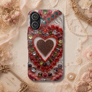 Op de afbeelding: Een smartphonehoesje met een centraal hart gevuld met kleine donkerbruine kristallen, omringd door een decoratieve rand van grotere rode, groene en heldere gefacetteerde kristallen op een witte achtergrond met kantpatroon.