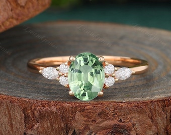 May include: A rose gold engagement ring with an oval green gemstone surrounded by smaller diamonds. The ring is set on a wooden surface.